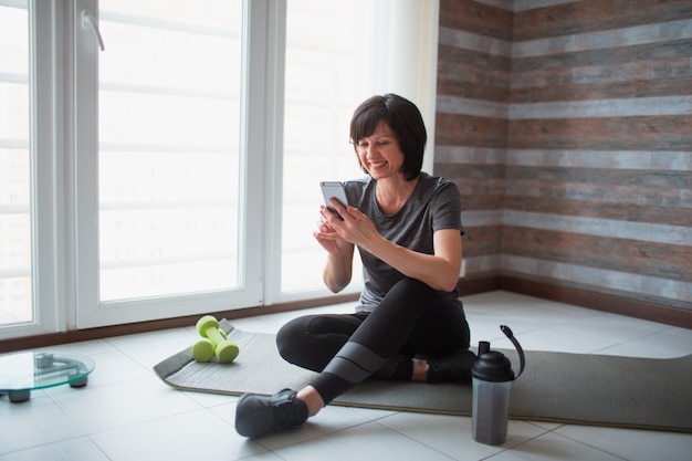 De volwassen geschikte slanke vrouw heeft thuis training. De vrouwelijke persoon zit op yogamat en gebruikt smatphone met handen. Ontspan en rust pauze of pauze tijdens het sporten.