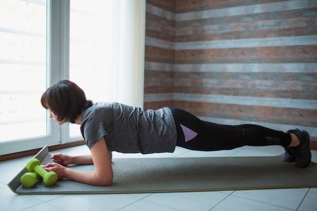 De volwassen geschikte slanke vrouw heeft thuis training. Ga op de halve plank staan en kijk vooruit. Rug en hele lichaam strekken. Goedgebouwde slanke volwassen vrouwentraining.