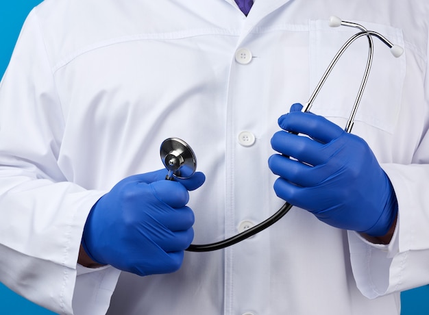 De volwassen mannelijke arts in een witte medische laag bevindt zich en houdt een zwarte stethoscoop op een blauwe achtergrond