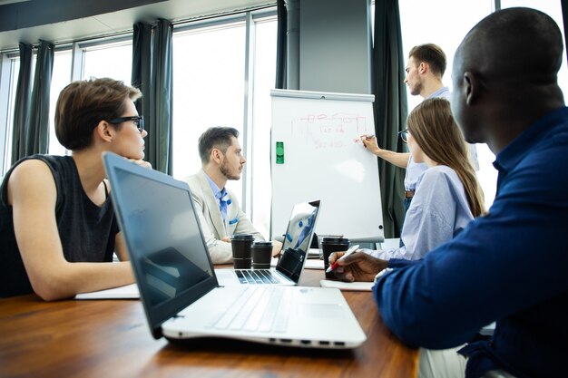 De voortgang van het bedrijf bespreken. Zelfverzekerde jonge man permanent in de buurt van whiteboard en grafiek te wijzen terwijl zijn collega's aan het bureau zitten.