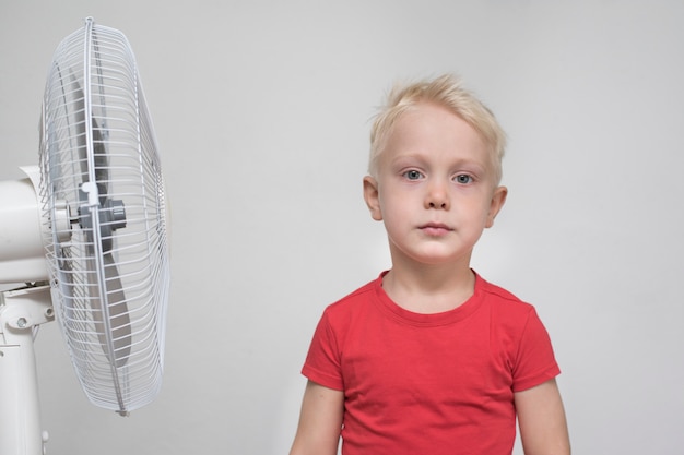 De vrij blonde jongen in rood overhemd bevindt zich dichtbij een ventilator.