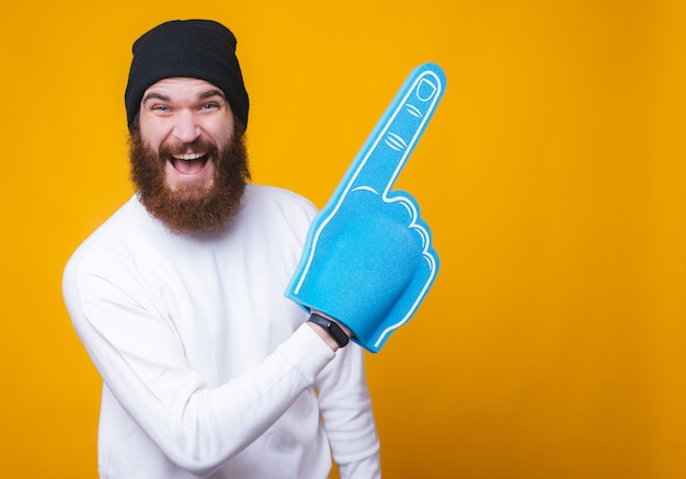 De vrolijke jonge gebaarde mens lacht en richt op een exemplaarruimte met een grote ventilatorhandschoen op gele muur.