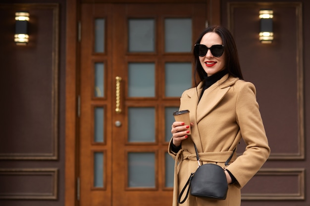 De vrolijke jonge vrouw die beige laag, zwarte sweater dragen, heeft handtas en zonnebril, drinkend afhaalkoffie in document kop, stelt glimlachend tegen de stadsbouw, wachtende partner voor vergadering.