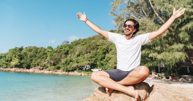 Foto de vrolijke latijnse jonge mens in witte t-shirt en zonnebril met uitgespreide wapens opende zitting bij het tropische strand, geluk, vrijheid van vakantie en reisconcept.