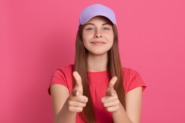 De vrolijke mooie vrouw die met tevreden gelaatsuitdrukking richten, heeft tevreden blik, dragend rode t-shirt en honkbalpet, geïsoleerd stellen over roze muur.