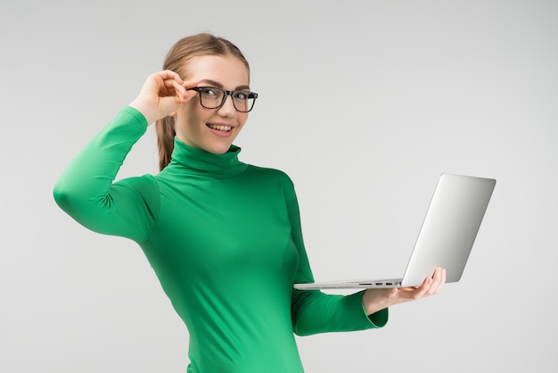 De vrolijke vrouw in glazen werkt aan laptop die het in haar handen houden terwijl status tegen op de witte achtergrond. Kijkt naar de camera