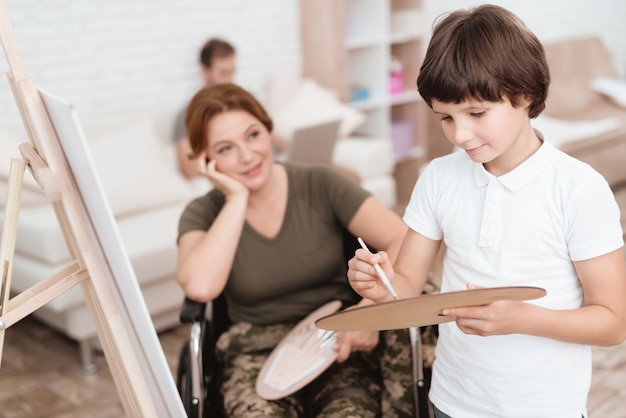 De vrouw in een rolstoel let op haar zoonsverven op canvas.