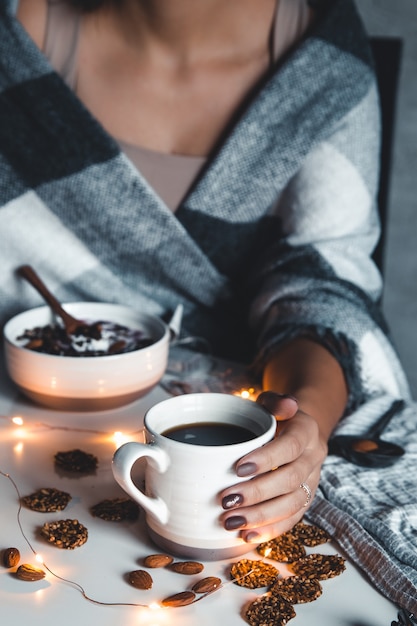 De vrouw is gewikkeld in een deken en heeft een kopje koffie in haar handen. Wintercomfort, slingers, kerstsfeer.