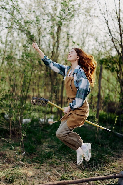 De vrouw springt amusant met haar hark als een heks op een bezemsteel in haar groene tuin tussen de bomen en glimlacht Het concept van alledaagse levensstijl en vreugde in het moment