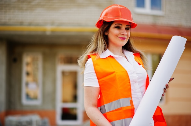 De vrouw van de ingenieursbouwer in eenvormig vest en oranje beschermend helm houdt handelspapier tegen de nieuwe bouw