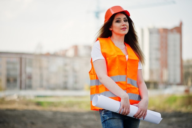 De vrouw van de ingenieursbouwer in eenvormig vest en oranje beschermende helm houdt handelspapier tegen nieuwe gebouwen met kraan.