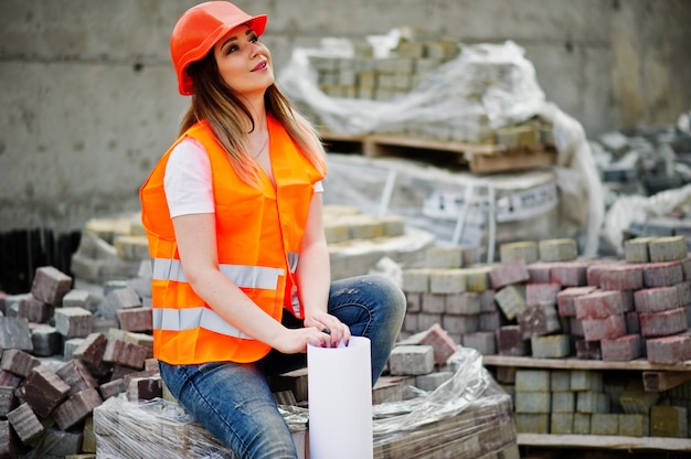 De vrouw van de ingenieursbouwer in eenvormig vest en oranje beschermende helm houdt het document van het bedrijfslay-outplan zitting op bestrating.
