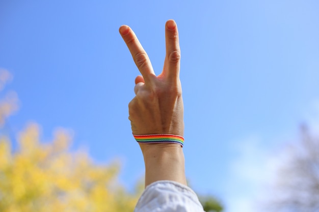 De vrouwelijke hand met regenboogarmband toont overwinningsteken op de blauwe hemel backgroung. LGBT-concept