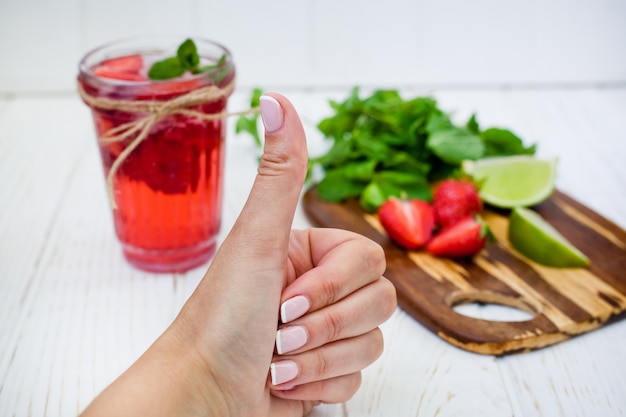 De vrouwelijke hand toont een goed teken op een achtergrond van aardbeidrank in een glas met limoen en munt