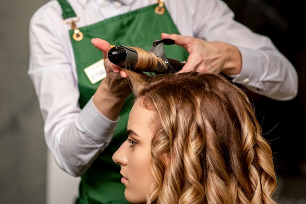 De vrouwelijke kapper krult haar voor een bruinharige jonge blanke vrouw in een schoonheidssalon