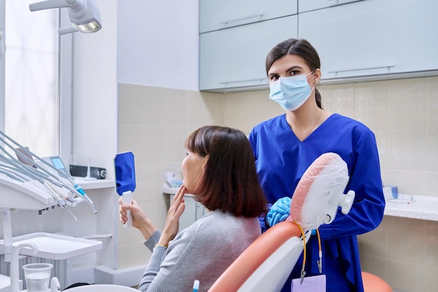 De vrouwelijke patiënt van het tandartspraktijk kijkt naar haar tanden in de spiegel