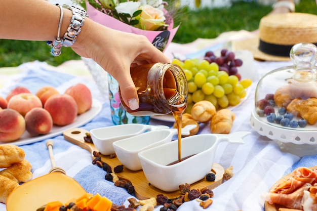 De vrouwenhand giet honing van transparante fles aan sauceboat, bij de achtergrond van de de zomerpicknick