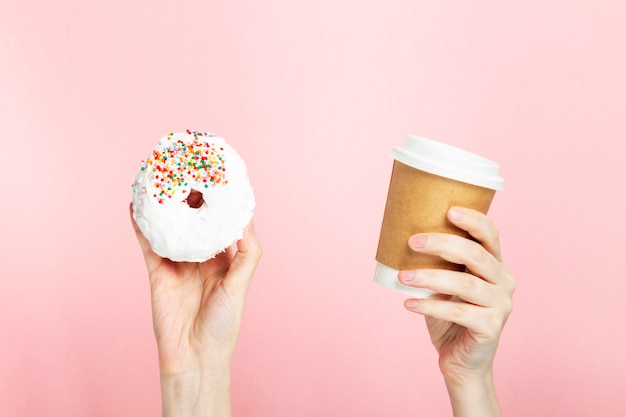 De vrouwenhand houdt document kop met koffie en doughnut met wit suikerglazuur en het veelkleurige bovenste laagje van het suikergebakje, roze achtergrond