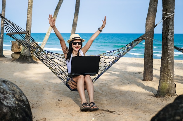 De vrouwenzitting van Azië op de hangmat dichtbij het strand en het gebruiken van laptop voor haar werk en het controleren van zaken tijdens haar vakantie.