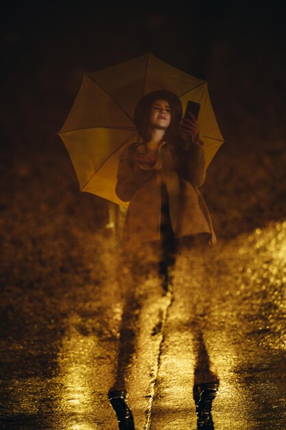 De weerspiegeling van een meisje in een regenvijver terwijl ze een smartphone gebruikt en een gele paraplu vasthoudt