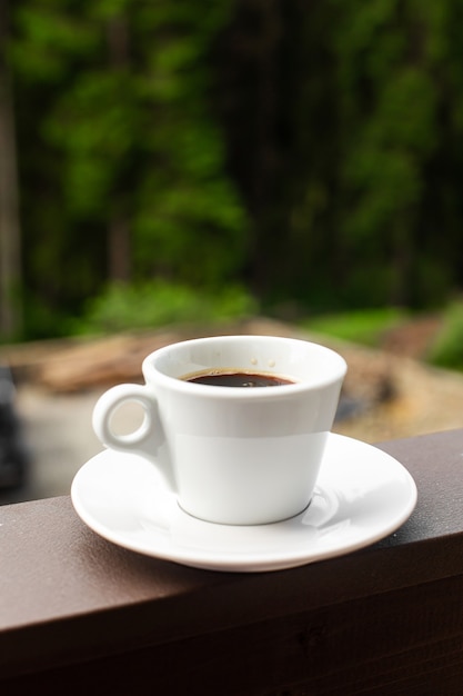 De witte mok donkere hete koffie in de ochtend op houten hek met bos achtergrond.