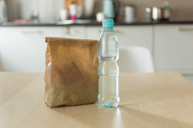 De zak van de pakpapierlunch en fles water op houten lijst over keukenachtergrond.