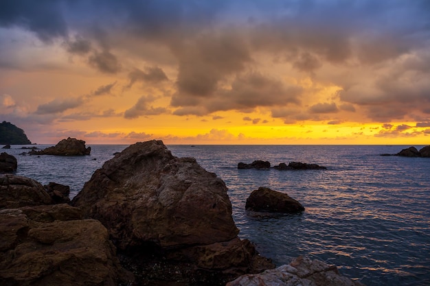 De zee en de rotsen en de stormachtige hemelbewolkte zonsondergang boven de zee in een rotsachtig strand met stenen en rood