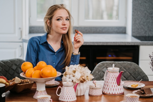 De zekere aantrekkelijke Kaukasische blondevrouw die blauw overhemd dragen houdt stuk van chockolate, drinkend thee terwijl het ontbijten in gezellige ruimte. Mensen thuis concept.