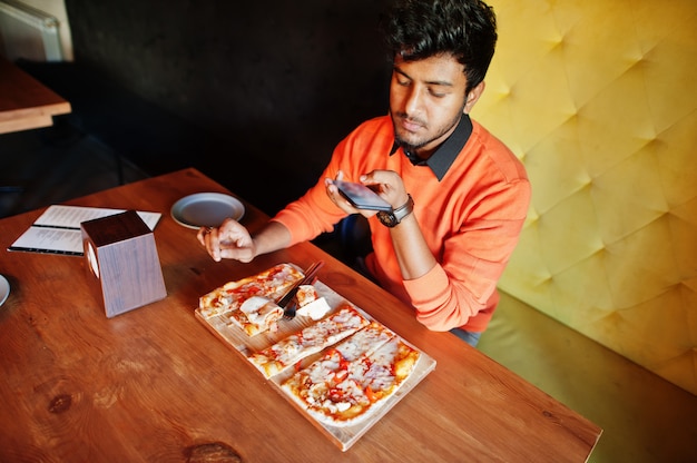 De zekere jonge Indische mens in oranje sweaterzitting bij pizzeria, eet pizza en maakt foto op zijn telefoon.