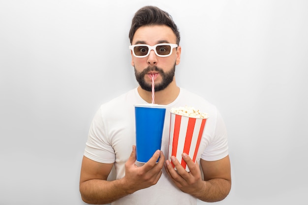 De zekere jonge mens kijkt door filmglazen. Hij houdt een glas cola nad popcorn in beide handen. Jonge man drinkt met rietje.