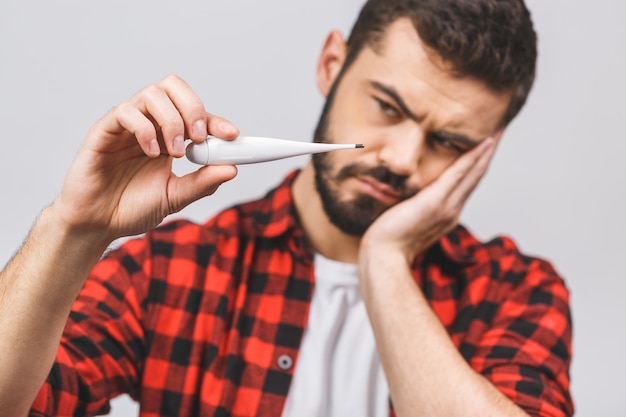 De zieke jonge mens bekijkt een thermometer die tegen witte achtergrond wordt geïsoleerd.