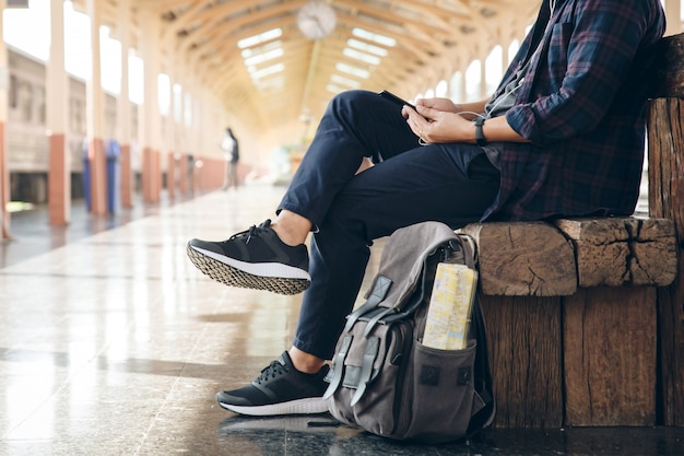 De zitting van de jonge mensenreiziger met het gebruiken van mobiele telefoon kiest waar te reizen en zak wachtend op trein bij station. Rugzak bij het station en het kijken op mobiele telefoon voor plan om te reizen.