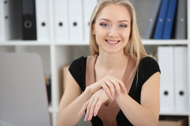 De zitting van het bedrijfsvrouwenblonde in het bureau bij een lijst, glimlachend en bekijkend de camera