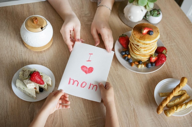 De zoon geeft zijn moeder een kaartje met moederdag Ontbijtverrassing van een kind Handen op tafel met pannenkoeken neem een kaartje