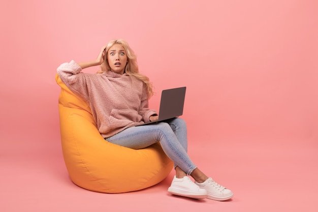 Deadline concept shocked young woman sitting in beanbag chair with laptop and grabbing her head pink