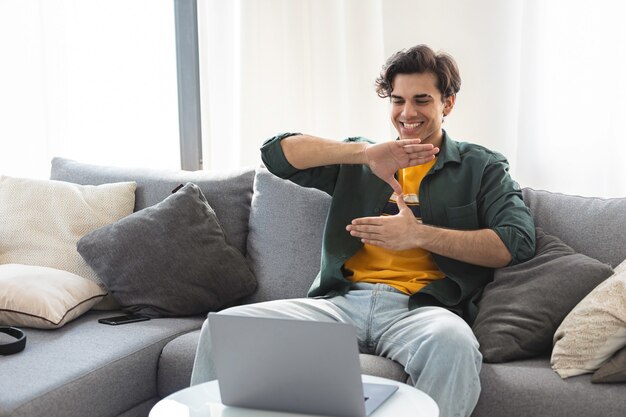 Photo deaf or hard hearing happy smiling young caucasian man uses sign language while video call using laptop while sitting on the couch at home