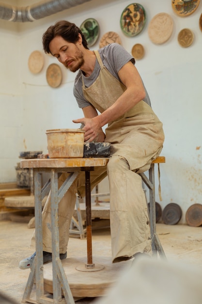 Dealing with clay. Bearded experienced man in grey apron sitting on high chair and intensely processing clay