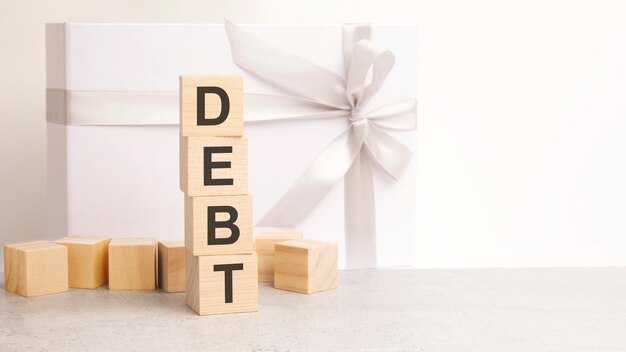 債務のテキストは、背景にある木製の立方体のピラミッドに配置されています。光沢のある白いリボンが付いた紙のギフトボックスです。
