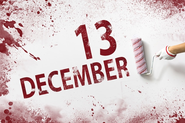 12月13日。月の13日目、カレンダーの日付。手は赤いペンキでローラーを保持し、白い背景にカレンダーの日付を書き込みます。冬の月、年の日の概念。