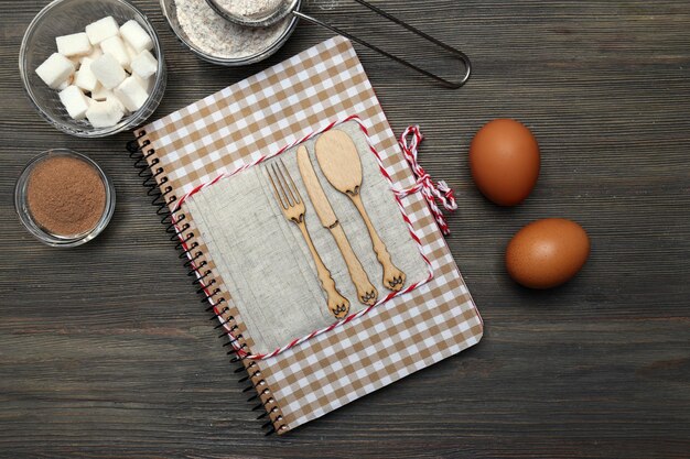 Decorated cookbook with ingredients for tasty cake on wooden background