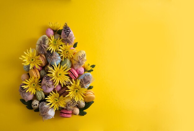 Decorated easter eggs in a composition with yellow flowers and macaroons on a yellow background