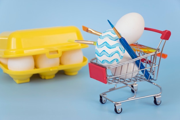 Decorated Easter eggs and paintbrushes in shopping cart with yellow egg tray filled with eggs on blue background.