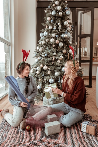 Decorated tree behind. Good-looking inspired ladies spending time in living room with gifts and paper for wrapping Christmas concept