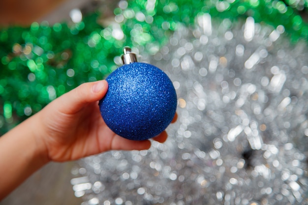 Decorating christmas tree at home. hand holding blue ball ornament close up on shiny background with colorful lights. space for text.