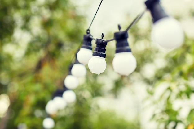 decoration, illumination, electricity, holidays and lightning concept - close up of electric bulb garland hanging in rainy summer garden