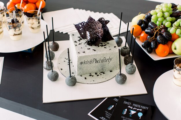 Decorations for birthday party. Stylish white and black decorated birthday cake, delicious desserts and fresh fruits on the black table. Birthday party concept, cake, desserts, candy bar