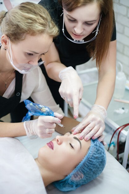 装飾美容コース。眉毛のマイクロブレードにタトゥーマシンを使用する方法を女性インターンに教える美容師。