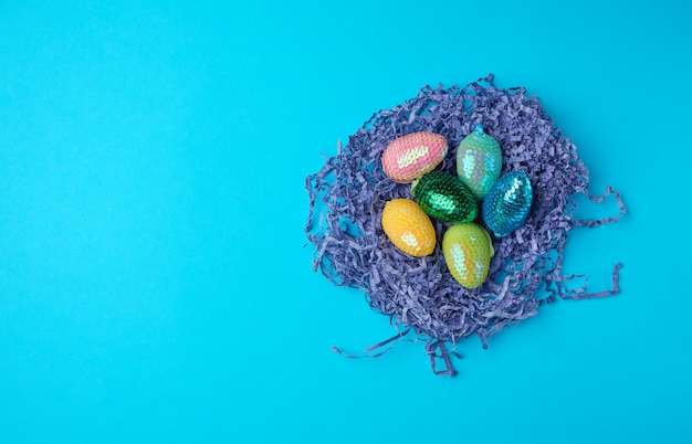 Decorative Easter eggs from sequins lie in a nest of strips of paper on a blue background