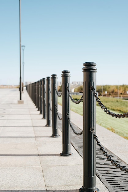 Фото Декоративный забор из черных стальных столбов с цепью на тротуаре в солнечный день