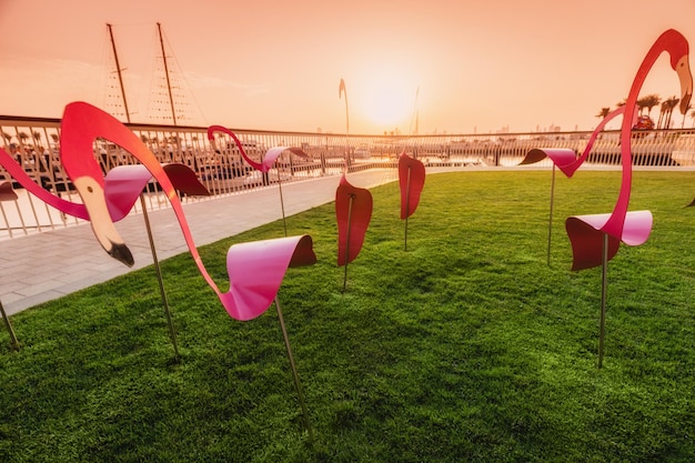 decorative figurines of pink flamingos on the boardwalk and white arch against the background of the bay with ships and yachts in the marina Creek Harbor in Dubai